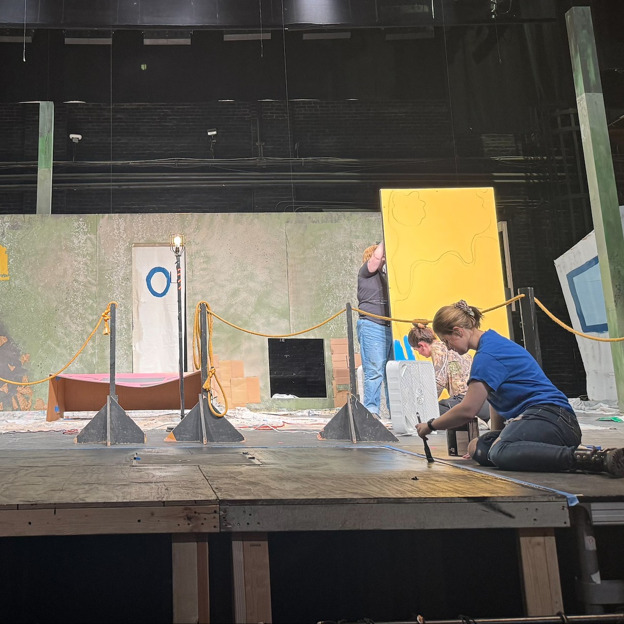 Several people painting various pieces of scenery on a theatre stage.
